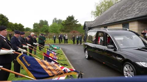 The image shows people on the left holding flags as a hearse carrying the coffin pulls up alongside the crematorium. 