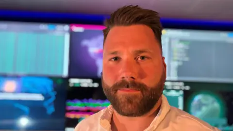 BBC/Caroline Bilton Headshot of a man with short brown hair and a brown beard and moustache wearing a white shirt. He is stood in front lots of monitors.