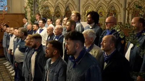 The choir stood singing to the audience at their annual Christmas concert inside Tyntesfield's chapel. Many are wearing denim jackets and are dancing to the carol.