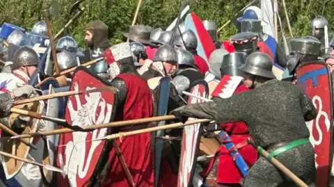 Mick Hurst A large number of men in medieval armour carrying spears and shields taking part in a mock battle with red shields