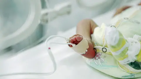 A stock image of a premature baby lying in an incubator in hospital, wearing a baby grow. It is a close up of the baby's legs with medical tubes coming from its foot.