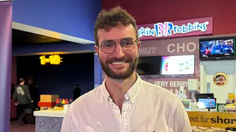 A man standing in front of a cinema check in desk with rows of bagged popcorn behind him and a Digital Jersey sign to his left.