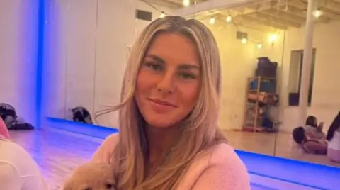 Ami Robertson sits in a Pilates studio. She is holding a dog on her lap and is smiling at the camera. She has long blonde hair.