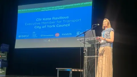 LDRS Councillor Kate Ravilious standing at a lectern and speaking onstage, against a black backdrop with a projector screen displaying her name. She wears a blue top patterned with white flowers and baggy beige trousers.