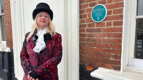 Ghost Tour operator Nicky Proctor wears a patterned black and red jacket and a top hat. She has blonde hair and red lipstick. She is standing outside a historic brick building with a blue plaque on it.