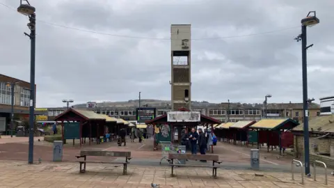 Three rows of fixed metal market stalls with a concrete clock tower in the background.