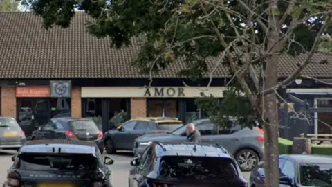 Retail units can be seen including one saying Amor in capital letters, with a cream coloured background and glass windows at the front. A car park is at the front of the building.
