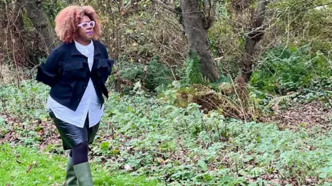 Errollyn Warren walks through the woods in her green boots. She wears a black skirt, black tights, a light blue shirt and a navy blue jacket. 