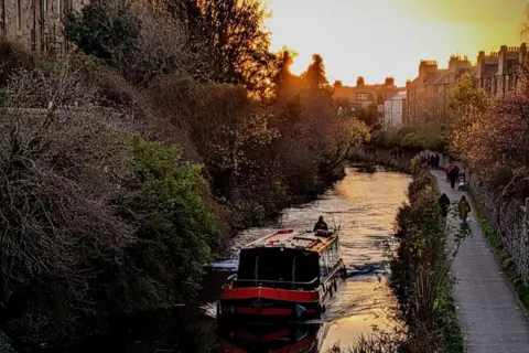 Vincent Maguire A narrowboat glides along a tree-lined canal as the sun sets behind rooftops.
