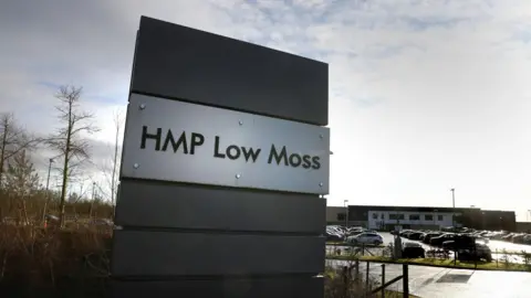 HMP Low Moss sign at its entrance with buildings and a car park full of cars in the background 