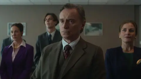 Netflix Robert Carlyle as Sam Hagen. He is stood in a suit with other actors playing members of the council in a drab office room