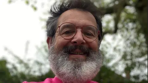 Charles Bain Smith A close up selfie of Charles wearing a pink fundraising t-shirt, he has red glasses and a bushy grey beard