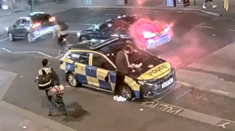 A man can be seen leaning over a police car with a takeaway tray placed on the bonnet. 