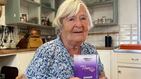 Genette sits at a wooden table in a homely kitchen, holding a purple “Make a Difference Awards – Winner” trophy. Behind her are open shelves, kitchen utensils, and a traditional stone feature above the cooker.