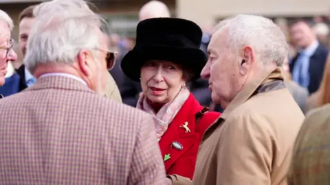 Mike Egerton/PA Princess Anne is in the centre of the photo. She is wearing a read coat covered in broaches. She has pearl earrings, a pink scarf and a fluffy black hat.