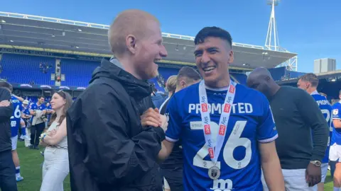 Connor Bennett/BBC Ed Sheeran is standing on a football pitch next to a football player who is on the right. Sheeran has short ginger hair and beard and is wearing a black coat. The football player - Marcelino Nunez - has short brown hair and is wearing a white and blue football T-shirt with 26 on the front.