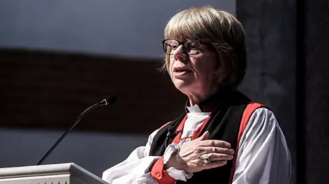 Dame Sarah Mullally, a woman with light-coloured hair in a short style with a fringe and glasses, stands at a lectern with a small microphone. She is wearing white, red and black religious clothing and has a large ring on her finger.