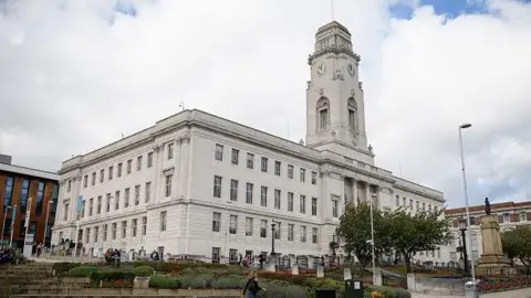 Getty Barnsley Town Hall situated along a street in an urban area. The building is constructed from light-coloured stone and has a grand, symmetrical design. A tall clock tower rises from the centre of the structure, with a clock face visible near the top. The tower is topped with decorative architectural details and stands prominently against the cloudy sky.