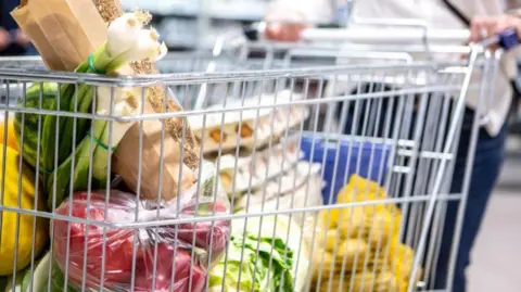 A supermarket trolley is filled with fresh produce, including spring onions, a bag of red peppers a yellow netted bag of potatoes and several large boxes of eggs.