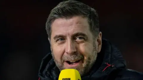 Getty Images Mark Chapman is smiling as he speaks into a BBC Sport microphone. He is wearing a black coat.