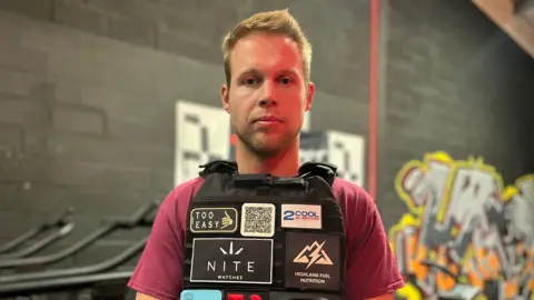 BBC A man wearing a burgundy t-shirt and a black vest is pictured stood in front of a black painted wall. 