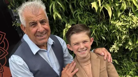 Marisa Billitteri Frankie wearing a light blue shirt and navy waistcoat holding the shoulders of Lucciano, who is wearing a camel-coloured coat and turtleneck jumper.