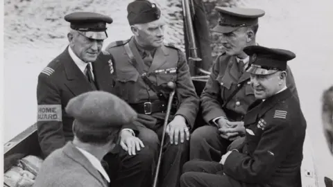 Royal Berkshire Archives Five men, four wearing uniforms of the British Armed Forces sit in a small boat on the water. The fifth man in the black and white image has his back to the camera and is wearing a jacket with a white shirt collar visible and a flat cap.