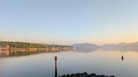 Roddy Forrest A calm body of water with two buoys, a rocky shore in the foreground, distant hills, and a clear sky at sunrise or sunset.