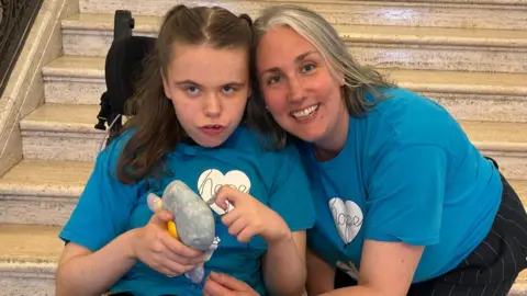 Alma White A woman in a wheelchair with dark hair and a woman with dark grey hair, they are both wearing blue and white tops.