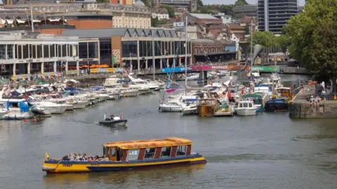 Paul Box A yellow Bristol Ferry boat in the foreground on the harbour with many other boats and buildings behind it