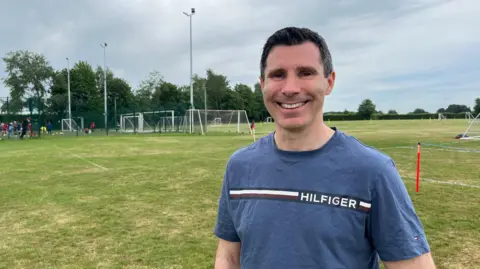 BBC A smiling man with black hair looking towards the camera. Behind him there playing fields with several football goals and white lines drawn on the ground. There is also a sports court with adults and children playing football wearing brightly coloured kits.