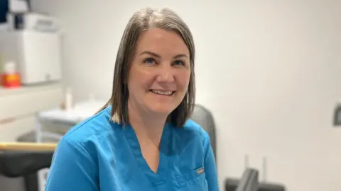 Federica Bedendo/BBC Gemma Hornsby-Lotfhouse is wearing a scrub-type blue top and is smiling. She has brown shoulder-length hair and blue eyes. The background is blurred and shows part of her treatment room.