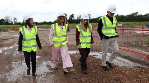 Reuters The image shows a group of four individuals—three women and one man—walking from across an outdoor, unpaved construction zone with puddles and gravel. All four are wearing personal protective equipment (PPE), including white hard hats and high-visibility yellow vests featuring a small logo on the left chest. They are dressed in smart-casual business attire underneath their safety gear. In the background, there is an open field, green trees under an overcast sky, and red metal construction barriers.