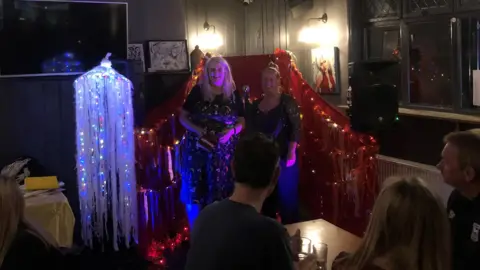 Jon Wright/BBC Ellen Clayton with poet and promotor Amy Wragg standing by a microphone on a stage in a pub. The stage is decorated with lights and flowing material. In the foreground there are people sitting at tables.