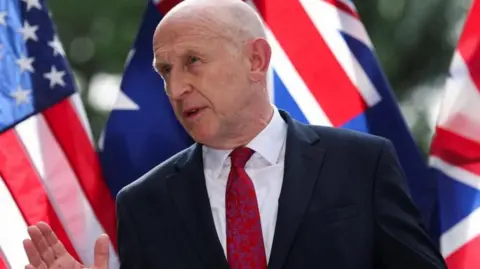 John Healey standing in front of the flags of the US, UK and Australia 