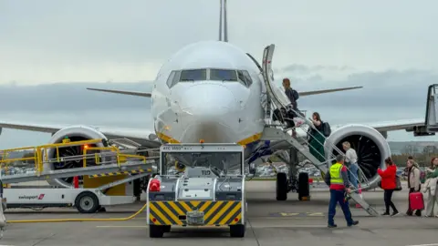 Getty Images A white and blue Ryanair plane on the runway at Bristol Airport. People are boarding it via the steps. 