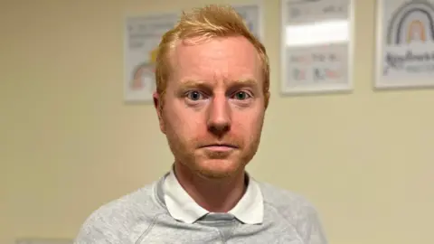 Gary stands in the middle of the image wearing a grey jumper with the collars of a white shirt visible. He has ginger hair and a ginger beard. 
