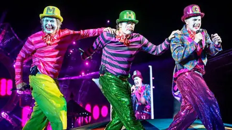 Getty Images The three men in pink and green circus outfits and face paint performing on stage in a row. They are all wearing bowler hats with the first letter of their name on. Gary Barlow holds a microphone up to his mouth, while Mark has his hand on Howard's shoulder, and Howard has his hand on Gary's shoulder. They all wear sequin trousers. 