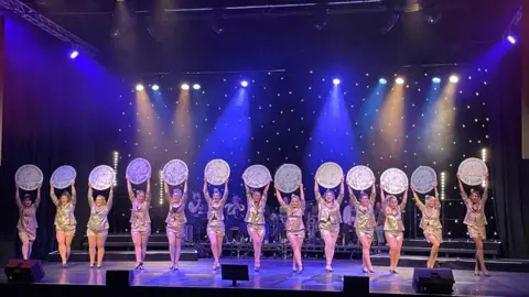 Hannah Ogden Women in golden dresses standing in a line on stage, holding round shields over their heads. 