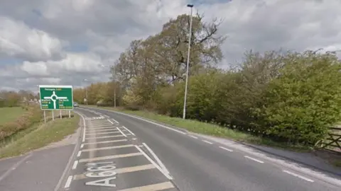 A stretch of the A661 sweeps to the left. In the distance is a large roundabout. The right-hand side of the road is flanked by trees and hedgerows.  