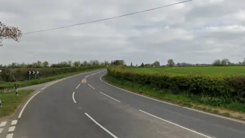 A curved section of rural road with the bend signage visible. There are hedgerows and fields on either side of the road. 