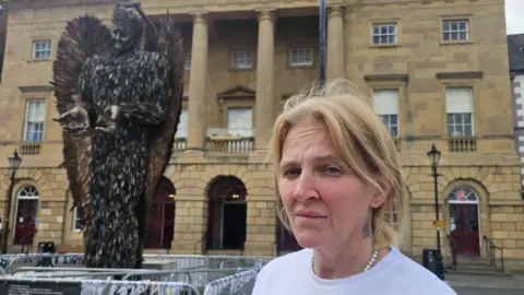 Adele Cook has blonde hair tied back and is looking at the camera. Behind her is a 27ft (8m) high statue of an angel made entirely from knives. 