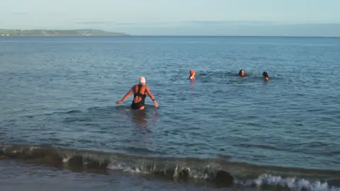 A woman in a black swim suit and white swim cap wades into the sea as a rolling wave crashes on the shore. Three other women are already mostly submerged in the water with their heads visible. Two of the women on the right have dark hair, the other is wearing an orange wooly hat and a red swim suit. Some land is visible to the left in the background showing another beach and some houses and fields.