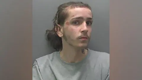 Cambridgshire Police Man with long hair tied in a pony tail with earing in one ear and a tattoo on the side of his neck. He has graze markings on his face and is wearing a grey sweatshirt.