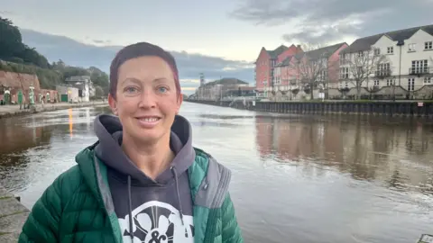 BBC Heather Baker from Saddles and Paddles wearing a mauve hoodie and a green padded jacket and standing next to the River Exe looking south.