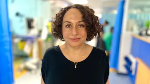 Dr Pathan is standing in a corridor in Picu. She has dark, jaw-length curly hair, is wearing a black, short-sleeved top and she is smiling. The background is blurred but shows a mix of blue cubicle curtains and machinery. 