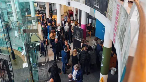 A crowd of people can be seen from above standing inside a building. There is a door to the left and columns through the middle. A large black sign is in front of the doors.