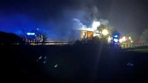 Bryan Stokes-Hazell A lorry can be seen on fire with a fire engine nearby. The lorry is slightly obscured by a tree and the image has been taken while it is dark.