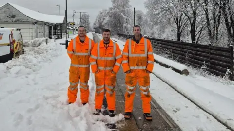 Network Rail Three men in hi-vis orange boiler suites standing on a snow-covered railway platform. 