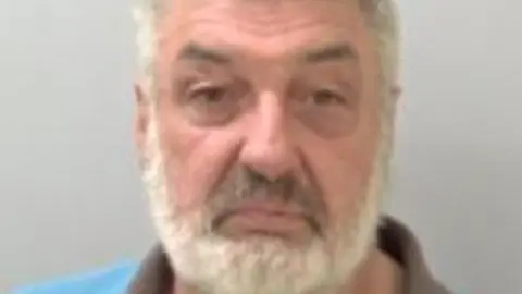 Hughes is wearing a blue top in his police mugshot and has a grey beard and hair.